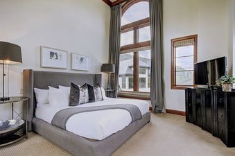A luxurious bedroom in the Ritz Carlton 2BR Valley View, featuring a plush king-size bed with white linens and gray bedding, two nightstands, a flat-screen TV on a dresser, floor-to-ceiling windows with views of the mountains, and gray curtains.