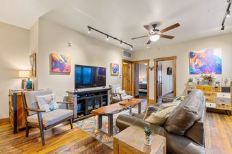 A cozy living room in Baker Loft 301 features a leather sofa, armchair, large TV, unique wooden coffee table, and colorful wildlife artworks, all set against rich hardwood floors.