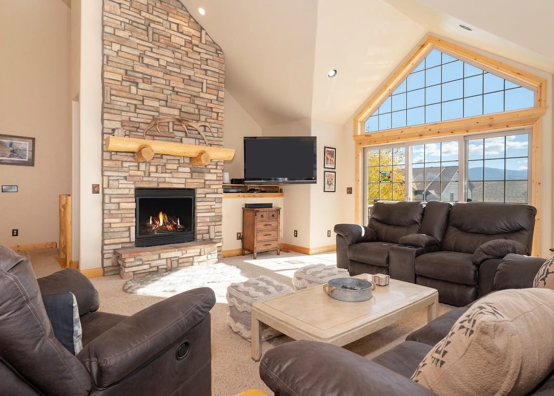 A spacious living room with a stone fireplace, comfortable sofas, and large windows offering a view of the mountains.