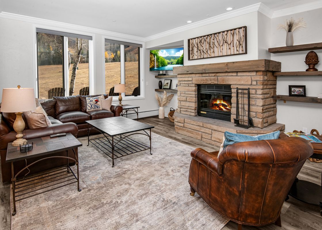 A warm and inviting living room at Highlands Slopeside in Beaver Creek, featuring a stone fireplace, comfortable leather seating, and large windows offering an autumn outdoor view.