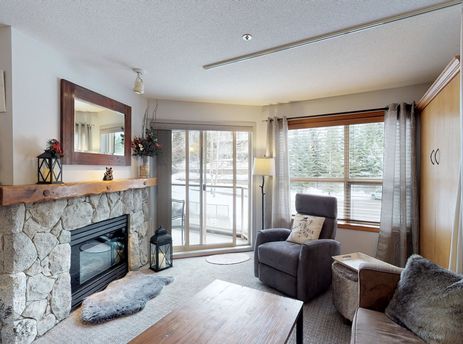 A cozy living room at Aspens on Blackcomb Unit 234 features a stone fireplace, comfortable seating, and a large window with a view of the snowy outdoors.