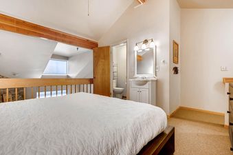The image shows a spacious bedroom with a white bed and a connected bathroom, featuring a vanity and toilet, under warm lighting and a wooden ceiling beam.