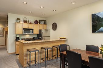 The kitchen and dining area at Villas at Snowmass Club 1515, featuring light wooden cabinets, stainless steel appliances, a breakfast bar, and a dining table with dark leather chairs.