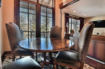 A round wooden dining table surrounded by four leather chairs with brass studding, set in front of large windows with blinds.