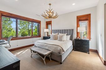 A primary bedroom at Blue Bird Vista at Hummingbird Lodge, featuring a king-sized bed with a gray headboard, flanked by dark nightstands and lamps, with a bench at the foot of the bed, and large windows offering views of lush trees and mountains.