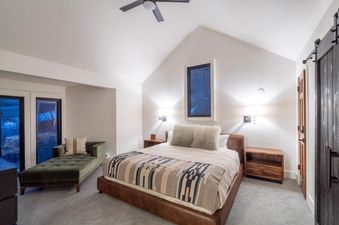 A master bedroom at Le Chamonix Unit A features a large bed, nightstands, and a chaise lounge, with vaulted ceilings and large windows providing natural light.