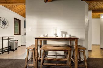 A cozy dining area at Cedar Creek Cabin featuring a wooden table with matching stools, set with white ceramic dishes and a central hurricane candle holder, under a vaulted wooden ceiling.