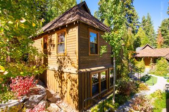 A two-story wooden cottage, Cedar Crest Eagle Cottage 8, with a gabled roof and numerous windows, is nestled among lush green trees and foliage under a clear blue sky, with a paved walkway leading to another cottage on the right.