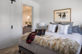 A cozy bedroom with a bed featuring a faux fur throw, a red plaid blanket, and a black handbag, with an open doorway leading to a bright bathroom.