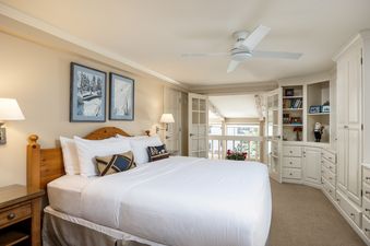 A serene bedroom at Montaneros 408 featuring a king-sized bed with white linens, two framed winter landscape artworks, a ceiling fan, and built-in shelving with decorative items, with French doors opening to an outdoor view.