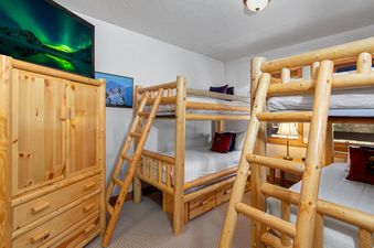 The bunk room at Town Pointe C302 features two wooden bunk beds, a large wooden cabinet with a TV displaying the Northern Lights, and a mountain art print.