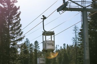 A gondola cabin from Winter Park ski resort, with the text 'Winter Park' on its side, is suspended in the air by cables against a bright sky, with tall pine trees and a colorful golden-leafed tree visible below and around it.
