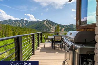The outdoor deck of Apex Manor features a grill and dining area, offering stunning mountain views under a bright sky.