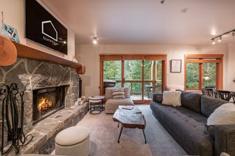 Cozy living room in Ravencrest 204 with a stone fireplace, comfy sofas, and large windows looking out to the forest.