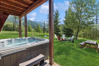 A hot tub on a covered patio with a view of mountains and a fire pit area in a grassy yard.