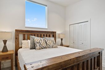 A cozy bedroom with a rustic wooden bed, white bedding, decorative pillows, nightstands with lamps, and a large window showing a blue sky.