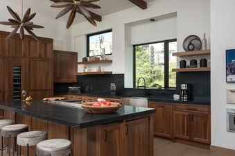 A modern kitchen with dark wood cabinets, black countertops, and a large island with bar stools, featuring unique light fixtures and large windows.