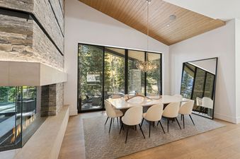 A modern dining room features a large rectangular dining table with eight chairs, an elegant chandelier, and a floor-to-ceiling fireplace, complemented by a large glass door leading to an outdoor patio with forest views.