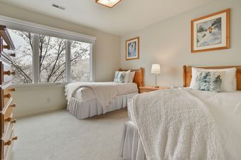 A cozy guest bedroom at Blue Bird on Norfolk with two twin beds, each covered in white bedding and a plush blanket, set against light-colored walls with framed winter art, and a large window offering a view of bare trees and a snowy landscape.