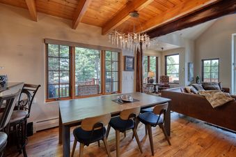 The image shows the dining area and living room of Moonlight Mountain 1 Hidden Trail, featuring a dining table, chairs, a chandelier, a leather sofa, and large windows with outdoor views.