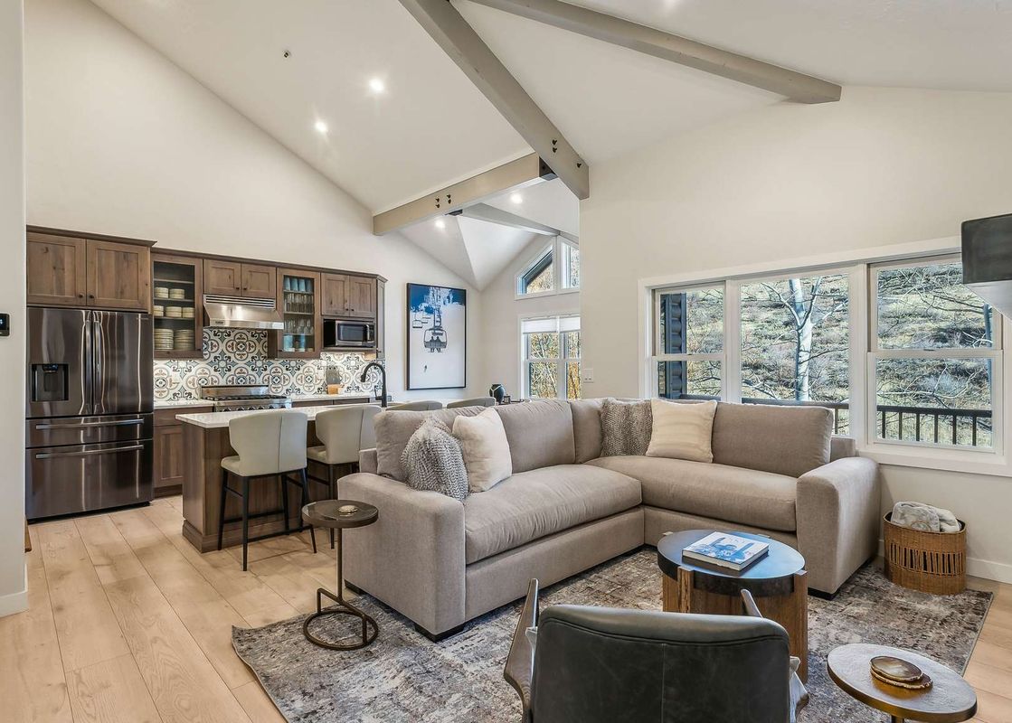 A modern open-concept living area with a kitchen, dining space, and lounge, featuring high vaulted ceilings and large windows showcasing an outdoor view.