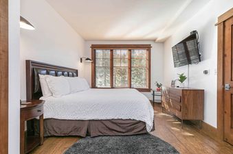 A bedroom with a white bed and brown headboard. The room has a window with blinds and a dresser.