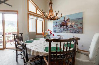The dining area at Strawberry Park 603 features a round wooden dining table with chairs, covered by a white tablecloth and green placemats, all illuminated by a unique antler chandelier. A large painting depicting cowboys and cattle adorns the wall, while