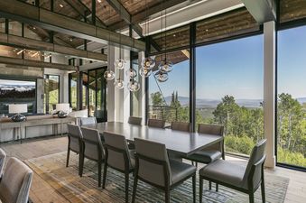 A luxurious open-plan dining area features a long dark wood dining table surrounded by light gray chairs, with contemporary glass pendant lights hanging above, and large windows offering a panoramic view of a lush green mountainous landscape.