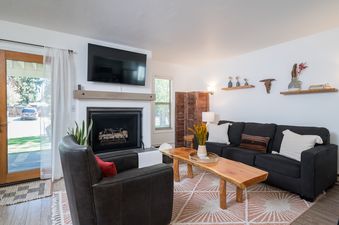 The living room at River Street Refugio features a dark gray sofa, a leather armchair, a rustic wooden coffee table on a geometric rug, and a fireplace with a mounted TV, all bathed in natural light.