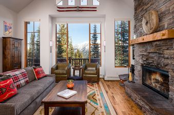 A living room with a fireplace, a large sectional sofa, two leather chairs, and a large window with views of the mountains.