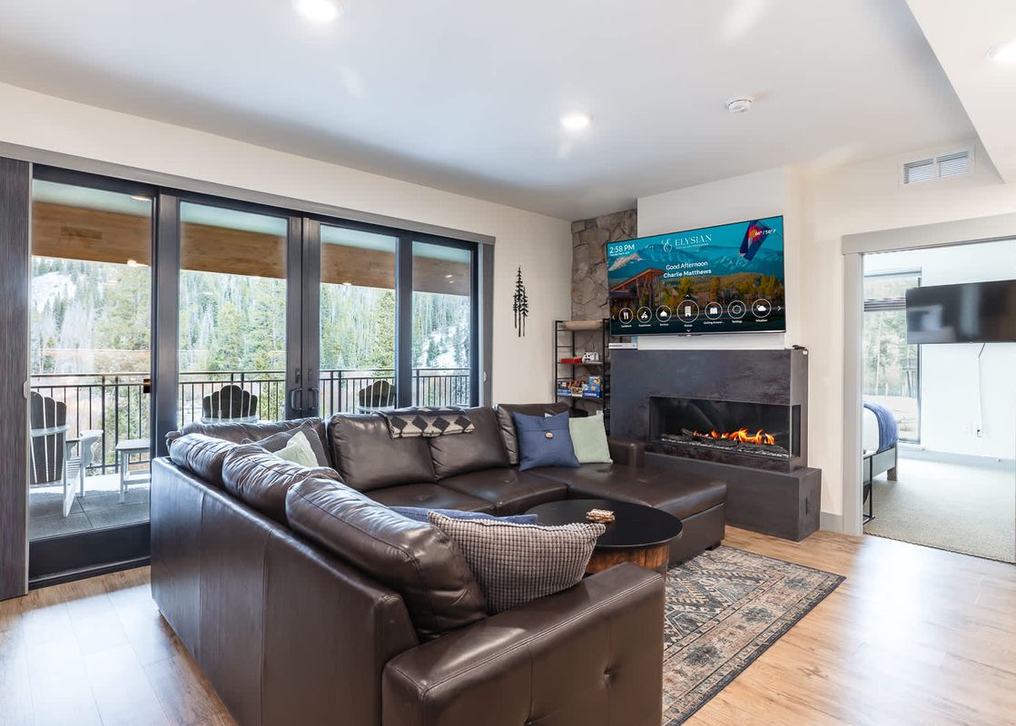 A luxurious living room in The Aerie features a large leather sectional sofa, a modern fireplace, a flat-screen TV, and sliding glass doors opening to a balcony with winter mountain views.
