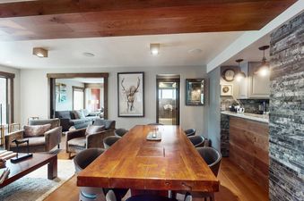 A long wooden dining table and chairs define the dining area, with a cozy living room visible beyond an open doorway, and a rustic-style kitchen with stone and wood accents to the right.