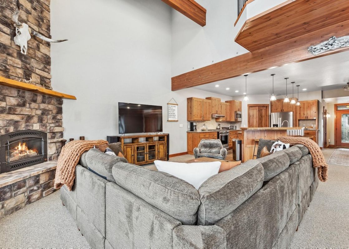 A cozy living room in the Chair View Chalet featuring a large L-shaped sofa, a stone fireplace with a fire, a flat-screen TV, and an open-concept kitchen with wooden cabinetry and a breakfast bar.
