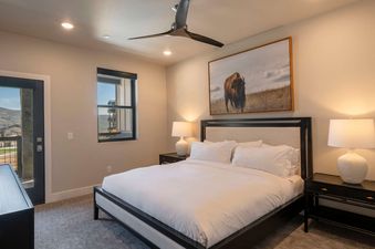 A spacious bedroom at The Retreat at Jordanelle features a large, comfortable bed with white linens, flanked by two black nightstands each holding a white lamp. A large framed picture of a bison hangs above the bed, and a ceiling fan is mounted overhead.
