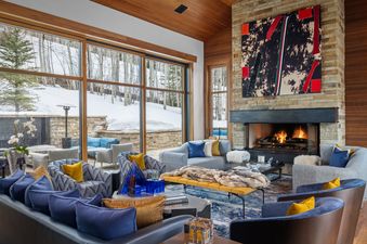 A luxurious living room with a stone fireplace, modern art, and large windows looking out onto a snowy, wooded landscape.