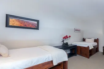 A bright and spacious bedroom at Lulu Luxe features two twin beds with white bedding, dark wood frames, and a shared console table adorned with a deer-shaped candle holder, all set against light gray walls and a beige carpet.