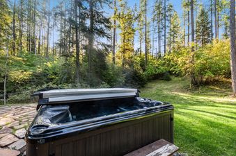 A hot tub with a partial cover sits on a stone patio surrounded by a green lawn and a lush forest under a sunny sky.