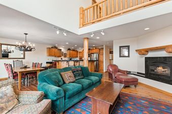 An expansive open-concept living space at Silvertip Ski Haus, showcasing a living room with a green sofa and fireplace, a dining area, and a kitchen under a loft.