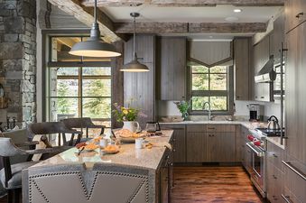 A large, well-lit kitchen with a spacious island, dark wood cabinetry, granite countertops, and modern stainless steel appliances, featuring views of the outdoors through large windows.