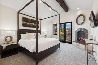 A luxurious primary bedroom with a dark wood four-poster bed, white bedding, an abstract painting, a stone fireplace, and large windows revealing an outdoor view.