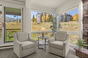 A cozy sitting area at Enclave 209 with two armchairs, a side table, and a lamp, offering a view of a mountain landscape with autumn trees and distant buildings through large windows.