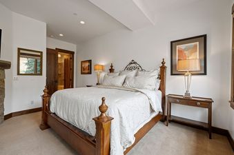 The luxurious primary bedroom at Juniper Landing 108 features a king-sized wooden bed with an ornate iron headboard, crisp white bedding, and elegant bedside lamps, complemented by a stone fireplace and a mirror reflecting an outdoor view.