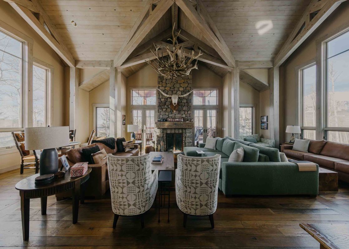 The living area at Powder Place on Lower Galloping Goose showcases high vaulted ceilings with exposed wooden beams, a grand stone fireplace with a longhorn skull mount, and large windows offering scenic winter mountain views.