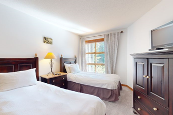 A bright bedroom at Aspens on Blackcomb Unit 332 features two twin beds with white linens, a nightstand with a lamp, and a large window with a view of lush greenery.
