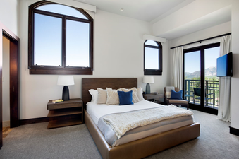 A king-sized bed with fluffy pillows and a soft throw blanket sits in the center of a bedroom with a mountain view from large windows and a sliding glass door.