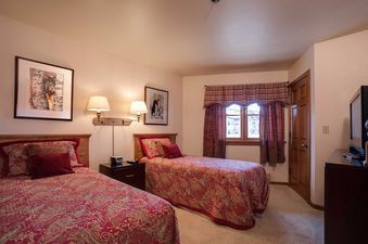 A cozy bedroom in Chateau Chamonix CX133 with two twin beds, each dressed in red and gold paisley bedding, a central nightstand, two wall sconces, framed artwork, and a window with patterned curtains.
