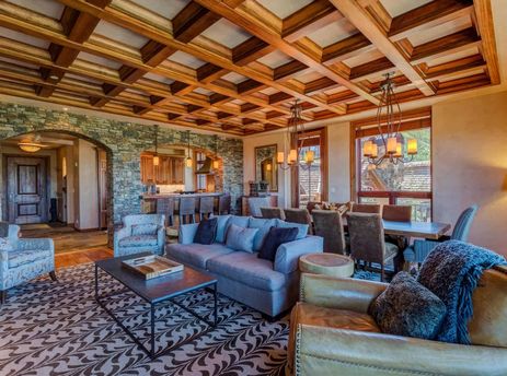 A luxurious and spacious great room with coffered wooden ceilings, a comfortable living area, an elegant dining room with chandeliers, and a glimpse of a gourmet kitchen through stone archways.