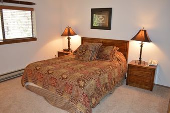The bedroom at Beaver Village 1633 features a queen-sized bed adorned with a colorful, patterned quilt, two bedside tables with lamps, and a window with blinds.
