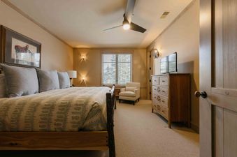A cozy bedroom with a wooden bed frame, a patterned duvet, and a ceiling fan, illuminated by natural light from a large window.