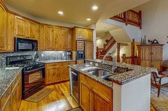 EagleRidge Townhomes 1512's kitchen is made up of dark wood cabinets, black appliances and a granite countertop. The kitchen overlooks a living room with a fireplace, a large flat-screen television and a couch. The kitchen has modern appliances